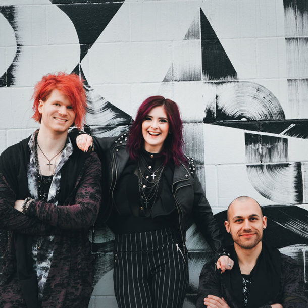 Photo of Trever Stobbe, Bethany Stobbe, and Wade Britz smiling together in front of black & white wall